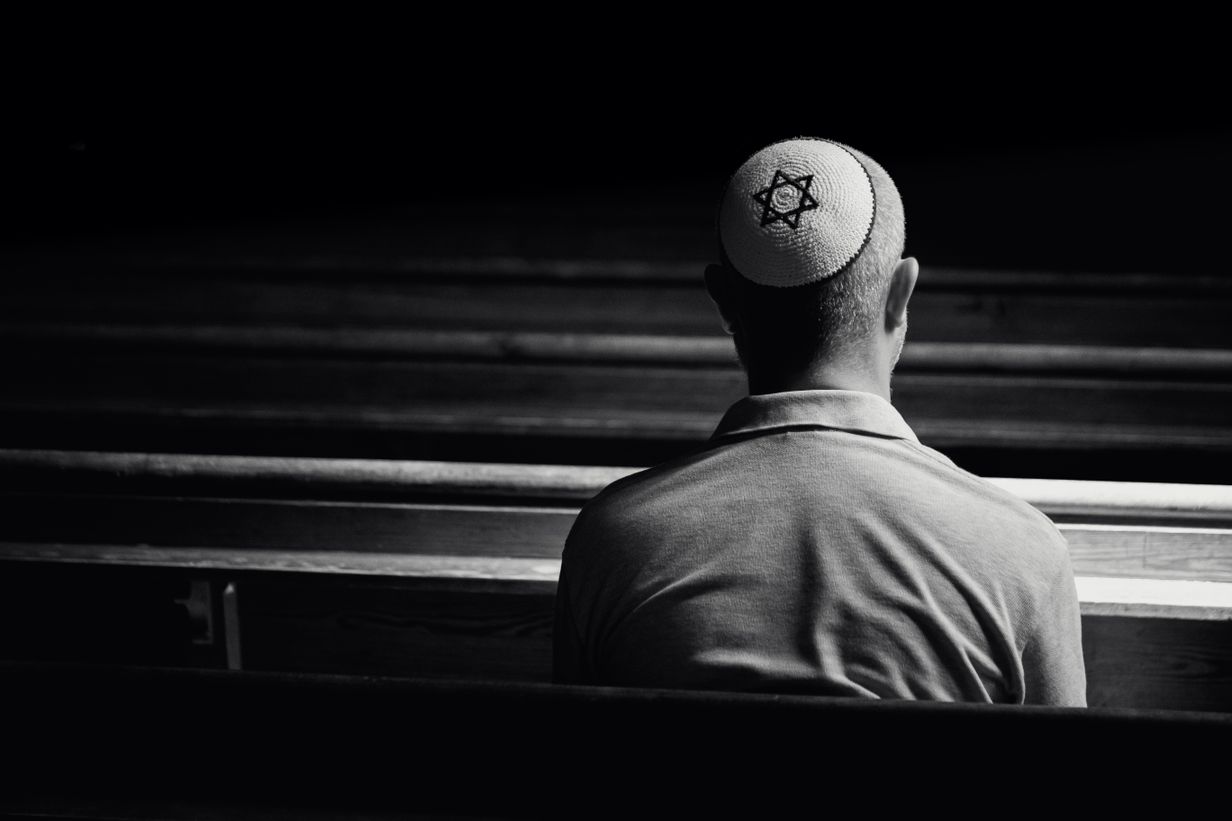 Monochrome image depicting a young caucasian Jewish adult man in his 30s inside a synagogue. He has his head bowed in prayer and he is wearing the traditional Jewish skull cap - otherwise known as a kippah or yarmulke - on his head. The man has a beard and the background of the synagogue is blurred out of focus. Horizontal color image with copy space.